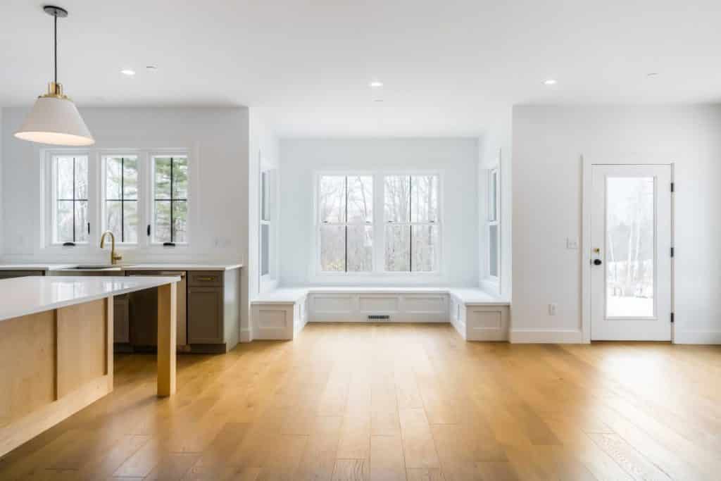 A large, mostly empty room with white walls. To the back of the room is a reading nook with windows on 3 sides and a window seat beneath. The kitchen is on the left side of the room; the rest is empty.