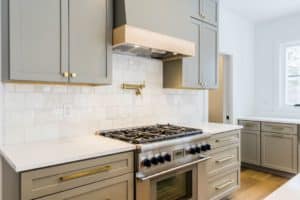 The range wall of a kitchen has light gray cabinets and a custom-made range hood with a band of neutral wood wrapped around the bottom. The backplash is made from textured white tiles.