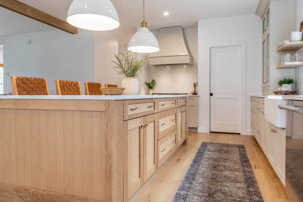 A generous island sits in the center of a boho chic kitchen. The base of the island is constructed of natural wood and contains frameless Shaker cabinets with gold hardware.