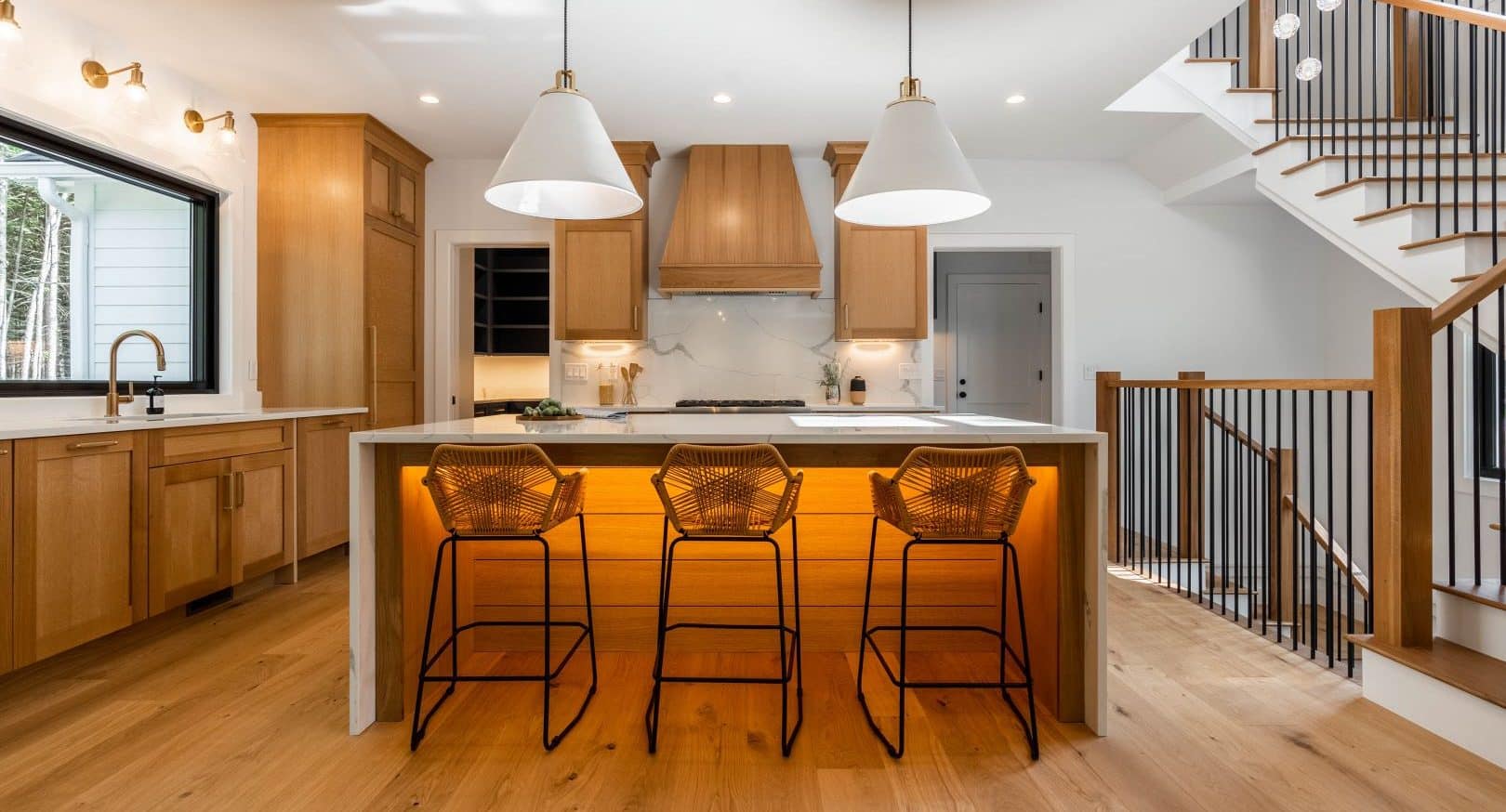 Broad view of a kitchen with a set of stairs to the right hand side of the viewer. The kitchen features natural wood flooring and cabinetry. In the center of the kitchen is an island with a waterfall quartz top, lit from below. Pulled up to the island are three cane bar stools with black metal bases.